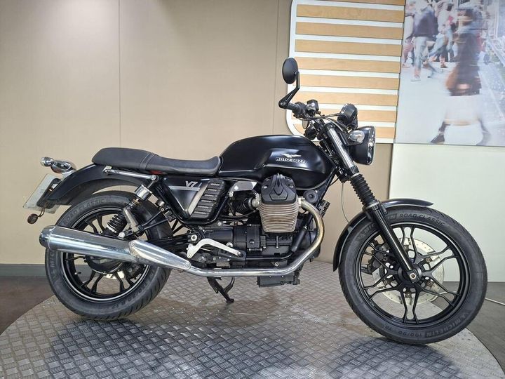 A black Moto Guzzi V7 motorcycle is displayed on a textured surface with a beige wall in the background. The bike features a retro design with chrome accents, a low saddle, and a prominent engine.