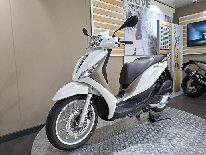 A white and brown scooter displayed on a textured platform in a showroom with a light-colored wall in the background.