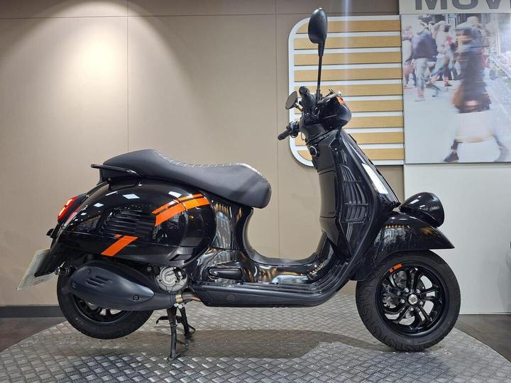 Vespa GTV 300 (2025) A black scooter with orange accents is displayed on a raised platform against a neutral background. The scooter features a rounded body, a comfortable seat, and sleek wheels.