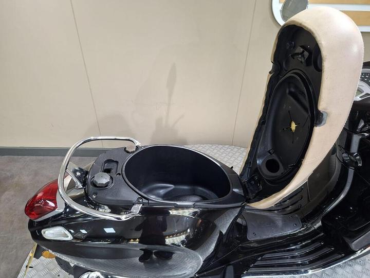 A close-up view of the seat of a black scooter with the seat raised, revealing a storage compartment underneath, featuring a round opening and a fuel cap.