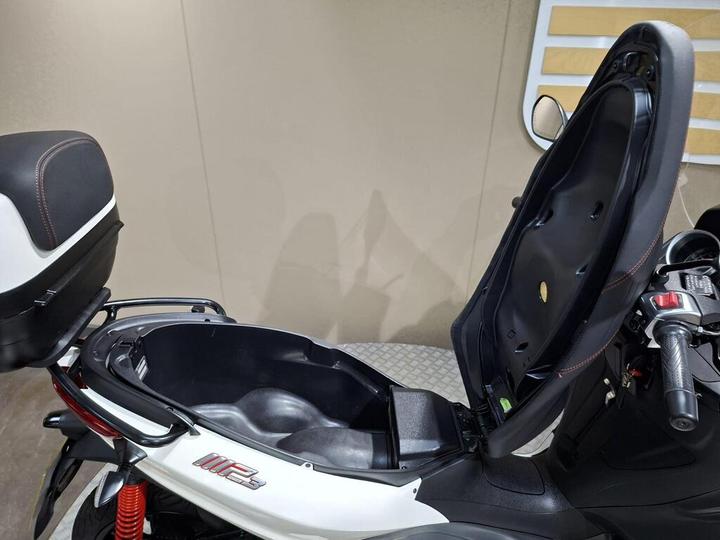 A close-up view of a scooter with the seat raised, revealing a spacious storage compartment underneath. The scooter features a white body with red accents, and the interior of the compartment is black. The background is a neutral color.