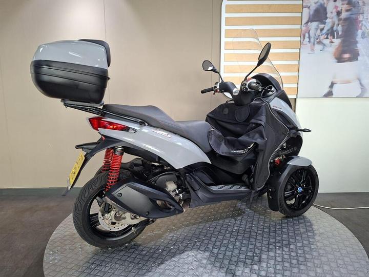 A gray three-wheeled scooter displayed on a circular platform, featuring a black top case and red shock absorbers. The scooter has a sleek design with a windshield and is positioned against a neutral background.