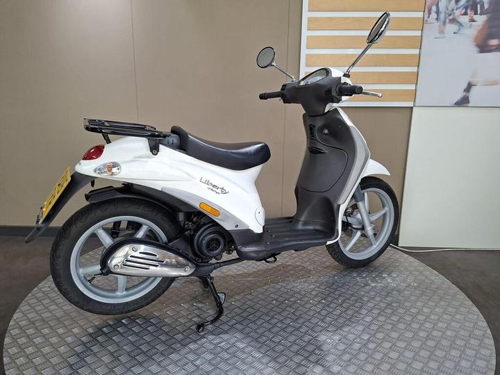 A white scooter with a black seat, parked on a circular metal platform. The scooter features a rear-mounted storage rack, a streamlined design, and mirrors on the handlebars. The background consists of a light-colored wall with a decorative panel.