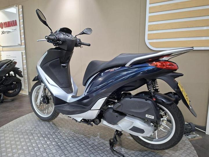 A sleek, dark blue scooter with a silver trim is displayed in a showroom, showcasing its stylish design and modern features. The background includes a Yamaha sign and another scooter partially visible.