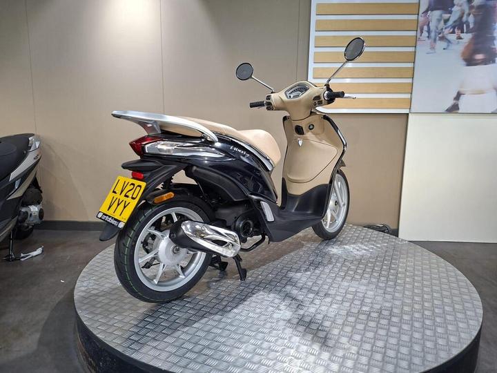 A black and beige scooter displayed on a circular metallic platform in a showroom, with a clear view of the rear and side details. The environment features neutral-colored walls and a minimalistic backdrop.
