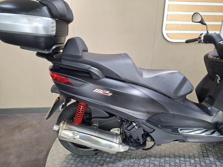 A close-up side view of a black scooter featuring a sleek design, a rear trunk, and a visible chrome exhaust. The suspension spring is red. The backdrop is a neutral-toned wall and patterned flooring.