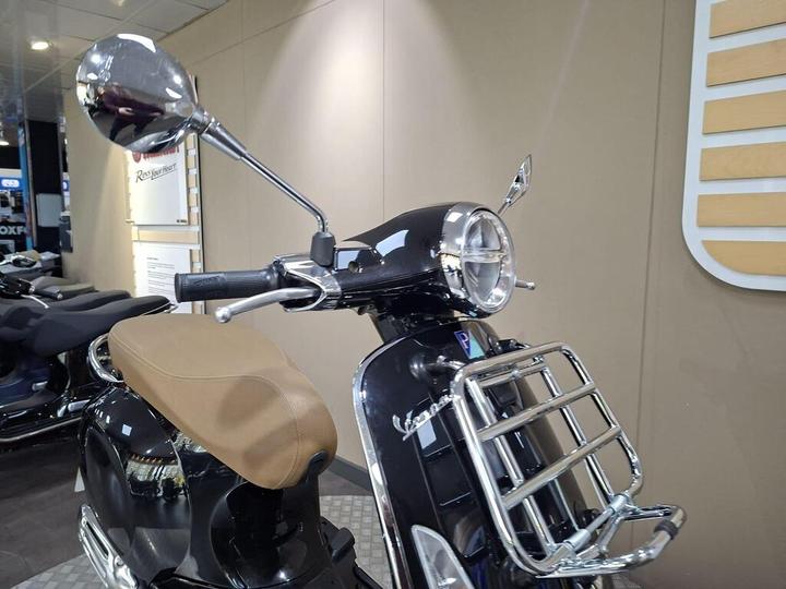 A close-up view of the front of a black scooter featuring a circular headlight, chrome mirror, and brown seat, displayed in a showroom with neutral-colored walls in the background.