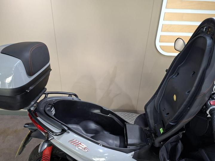 A close-up view of the storage compartment of a light gray scooter with a black and red seat back, partially opened to reveal its interior. The background features a light-colored wall and a wooden panel.