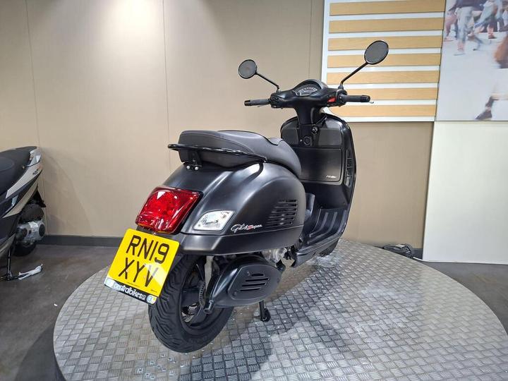 A black scooter parked on a circular metal platform, showing the rear and side views, with a visible license plate reading "RN19 XYV." The background features a beige wall with horizontal slats and a blurred image of people running.