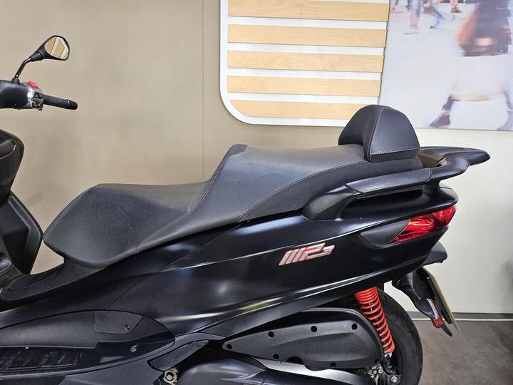 A black scooter parked indoors, showcasing its sleek design with a prominent seat, rear suspension in red, and a side view. The background features wooden paneling and a blurred image of people.