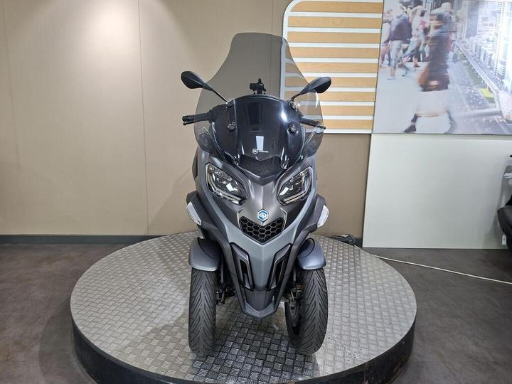 A front view of a gray three-wheeled scooter displayed on a circular platform, with a minimal background featuring a partial wall and an image of a busy street scene.