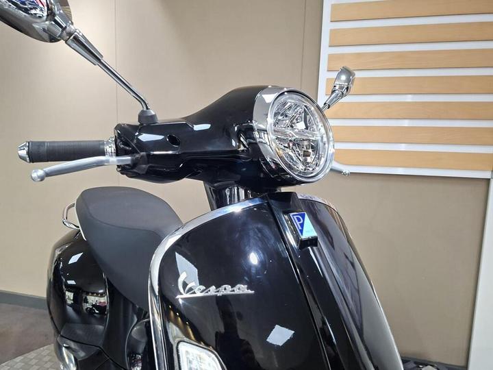 A close-up view of a black Vespa scooter's front, highlighting the handlebars, chrome details, and headlight, set against a neutral background.