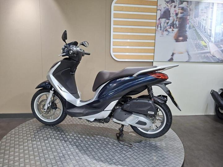 A blue and silver scooter is displayed on a circular silver platform against a beige wall, with a blurred image of a city street in the background.