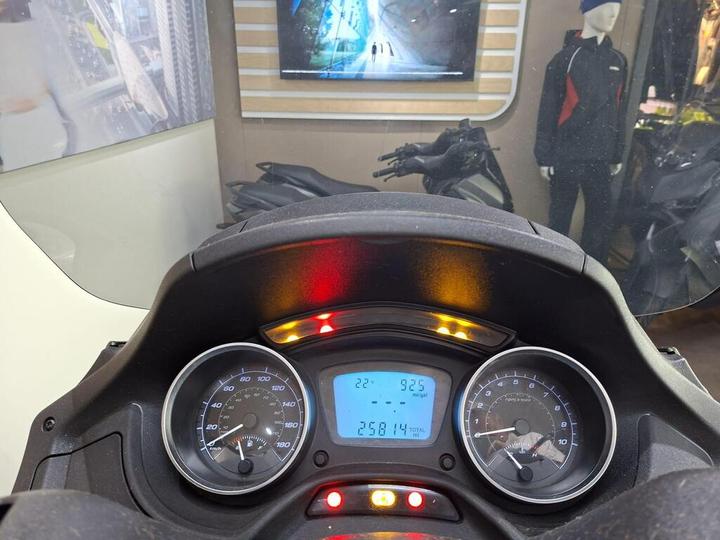 A close-up view of a motorcycle dashboard, displaying speed, mileage, and warning lights. The screen shows a speed of 92.5 miles per hour and a total mileage of 25814 miles, with various indicator lights illuminated. In the background, motorcycle displays and apparel are visible.