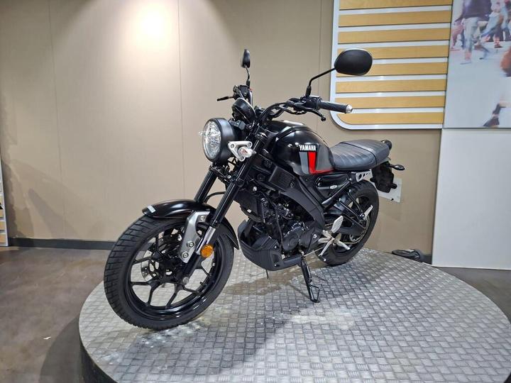 A black Yamaha motorcycle displayed on a circular metallic platform, featuring a minimalist design with a round headlight, sleek body, and textured seat. The background is neutral with a wooden panel and a partial image of people.