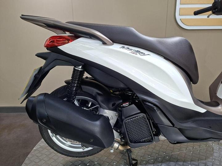 A close-up view of the rear section of a modern white scooter, featuring a sleek design, brown seat, and prominent exhaust system. The background is neutral, enhancing the scooter's polished appearance.