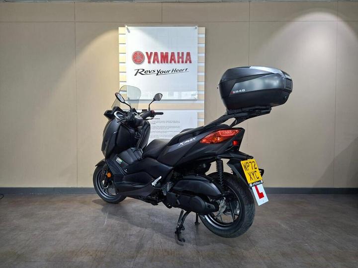 A black Yamaha scooter with a top box, positioned in a showroom with a Yamaha logo and slogan on the wall behind it. The scooter features a learner plate on the back and is displayed on a smooth floor.