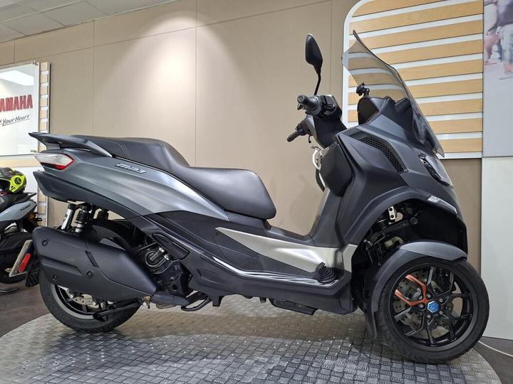 Piaggio MP3 530 HPE Exclusive 2023 A modern three-wheeled scooter in a showroom, featuring a sleek gray body, single seat, and prominent front windshield. The background includes a Yamaha sign and additional scooters.