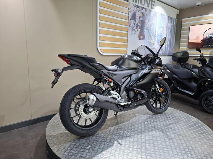 A sleek black motorcycle displayed in a showroom, featuring a modern design and prominent rearview mirrors, with a textured circular platform and a soft backdrop of beige walls and screens.