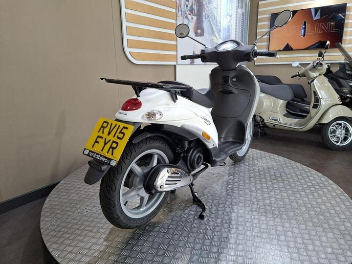 A white scooter displayed in a showroom, featuring a prominent license plate labeled RV15 FYR, with chrome details on the exhaust and a sleek design. In the background, another scooter is visible along with elements of the showroom decor.