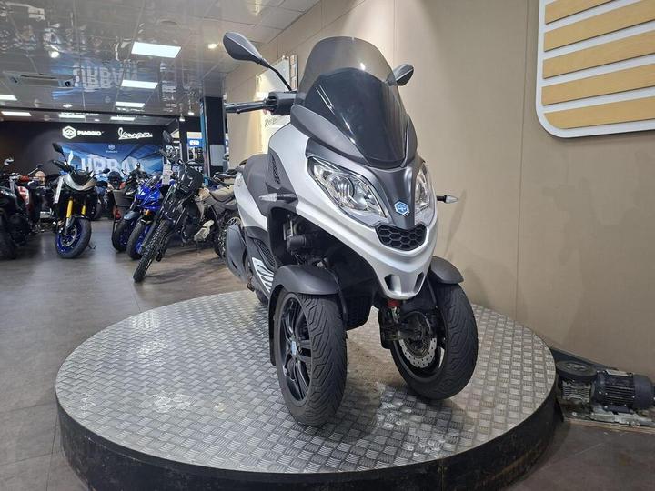 A silver three-wheeled scooter displayed on a rotating platform in a motorcycle showroom, with various motorcycles and sportbikes visible in the background.