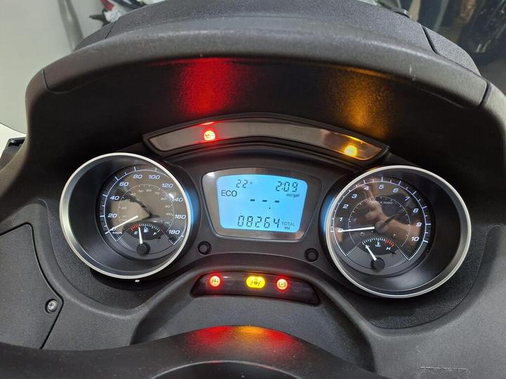 Close-up of a motorcycle dashboard displaying two speedometers, with the left showing speed and fuel level, and the right indicating RPM. A digital screen presents the temperature (22°C), time (2:09), and total mileage (8264 miles), accompanied by warning lights in red and yellow.