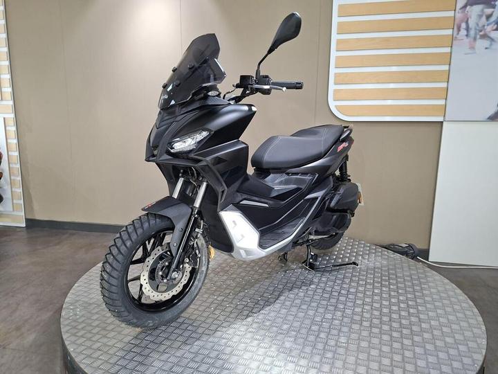 A black scooter with a protective windshield, showcased on a display platform with a textured surface, set against a neutral background.