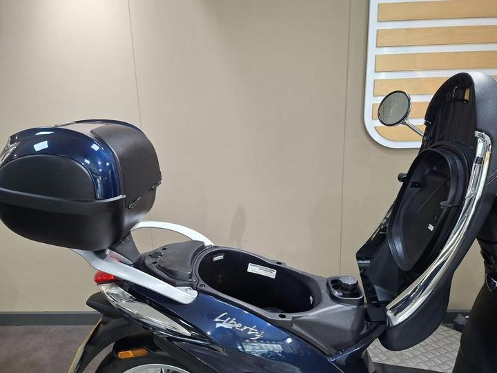 Close-up of a blue scooter with a black storage compartment and rear trunk, showcasing the open seat and storage area, set against a neutral backdrop.