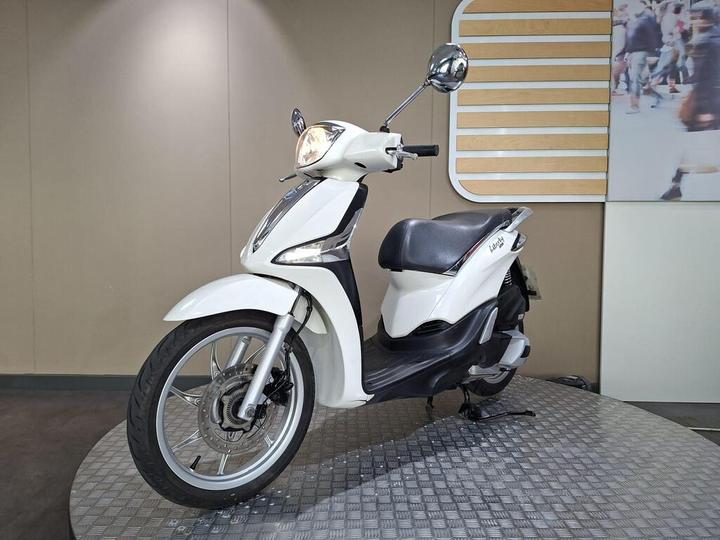 A white scooter is displayed on a circular platform against a neutral backdrop, showcasing its sleek design and features.