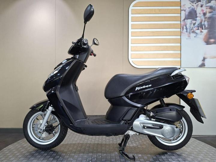 A black scooter, model Kisbee, shown from the side against a neutral background with a wooden and photo display. The scooter features a sleek design with a comfortable seat and a visible kickstand.