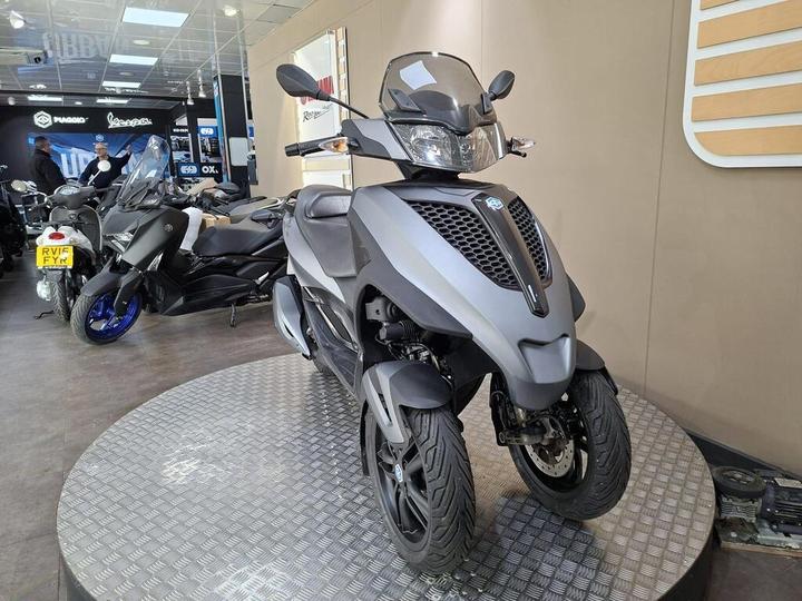A sleek gray three-wheeled scooter displayed on a circular platform in a motorcycle showroom, with other bikes visible in the background.