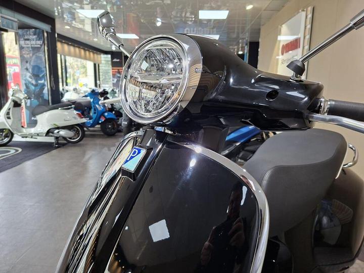 Close-up of a black scooter's front, featuring a round headlight and chrome detailing, with a blurred showroom background showcasing other scooters.