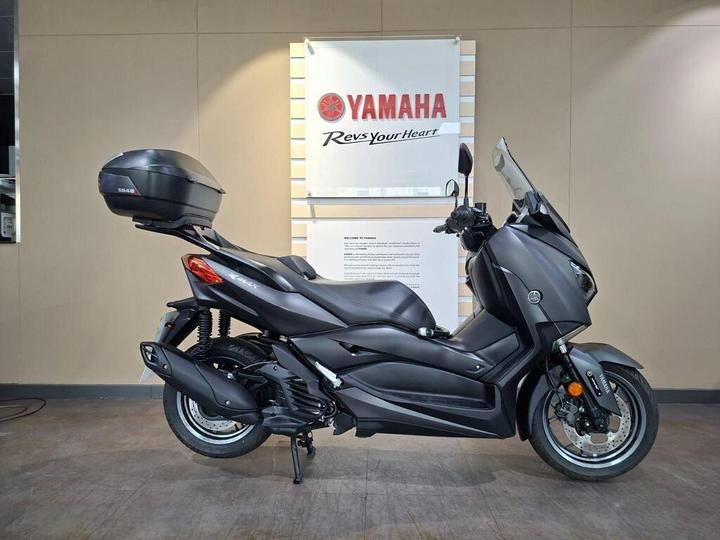 Yamaha XMAX 125 A dark gray Yamaha scooter parked indoors, featuring a rear storage box, visible brake disc, and sleek body design, with a Yamaha logo displayed on the wall behind it.