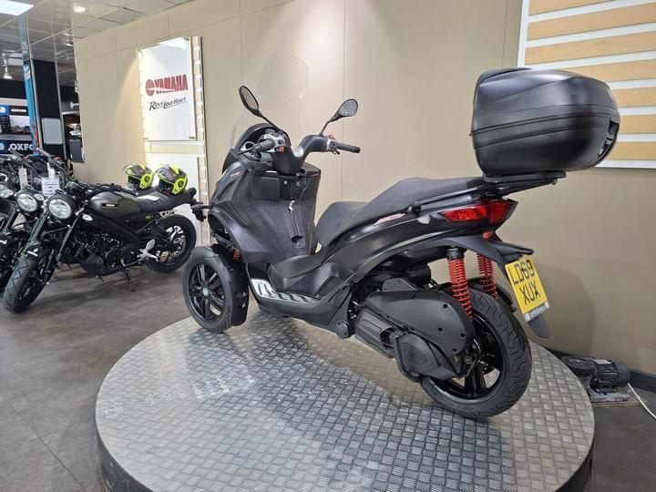 A black three-wheeled scooter displayed on a circular platform in a Yamaha dealership, with motorcycles and bright yellow helmets in the background.