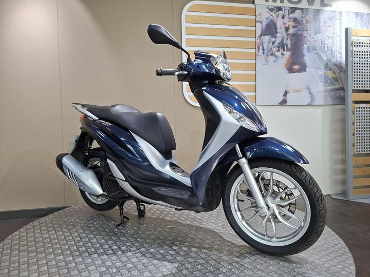 A blue and silver scooter displayed on a textured surface against a light-colored backdrop. The scooter features a sleek design with a large front headlight and modern alloy wheels. In the background, a blurred image of a bustling city scene is visible.