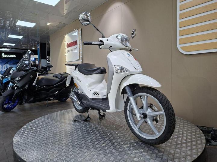 A white scooter is displayed on a circular platform in a showroom with a wooden and beige backdrop, alongside a black motorcycle.