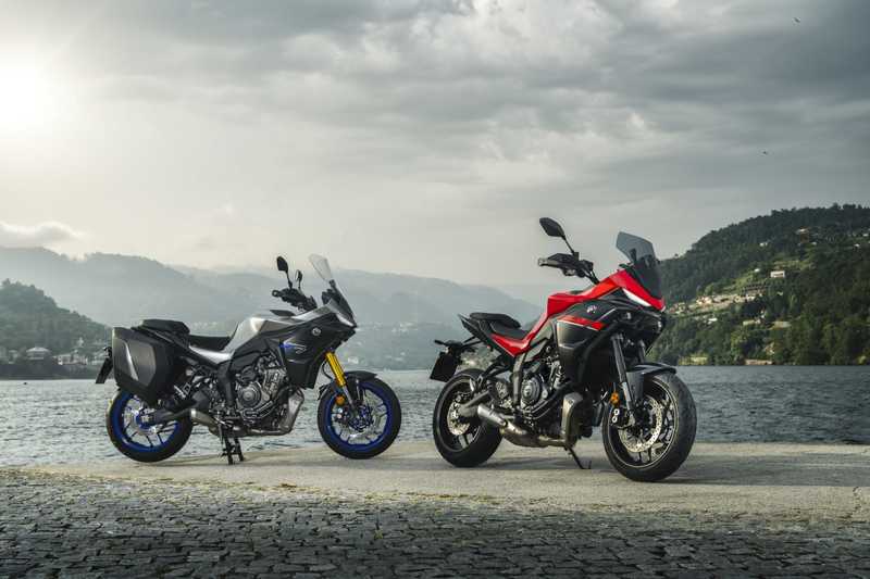 Two motorcycles are parked side by side on a cobblestone surface by a lake, with hills and a cloudy sky in the background. The motorcycle on the left is predominantly silver with blue accents, while the one on the right features a striking red and black design.