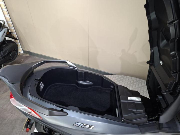 A top view of an open storage compartment on a grey scooter with a textured flooring surface in the background.