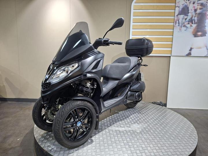 A black three-wheeled scooter displayed on a round, metal-patterned platform, featuring a windshield and a rear storage compartment, set against a soft-focus backdrop.