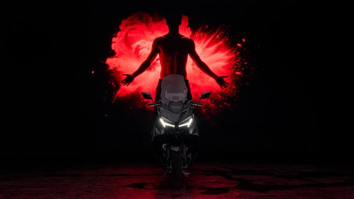 A sleek black motorcycle is positioned in the foreground, illuminated by dramatic lighting, with a striking red silhouette of a person with outstretched arms and an explosion effect in the background.