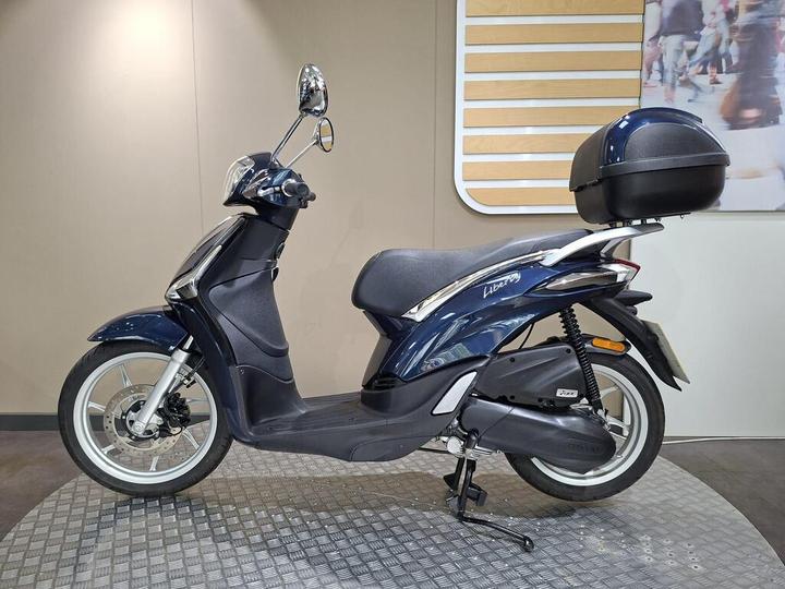 A blue scooter with a sleek design and a rear storage compartment, displayed on a textured surface against a light-colored wall.