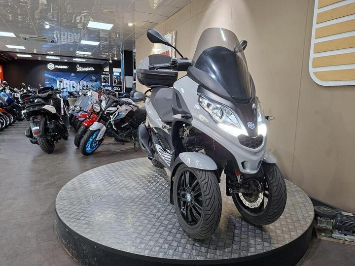 A gray three-wheeled scooter displayed on a circular platform inside a motorcycle showroom, surrounded by various other motorcycles. The showroom has modern lighting and branding from different motorcycle manufacturers in the background.