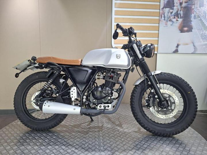 Mutt Akita 125 2024 A custom motorcycle with a silver fuel tank, brown leather seat, and rugged tires, displayed against a neutral backdrop.