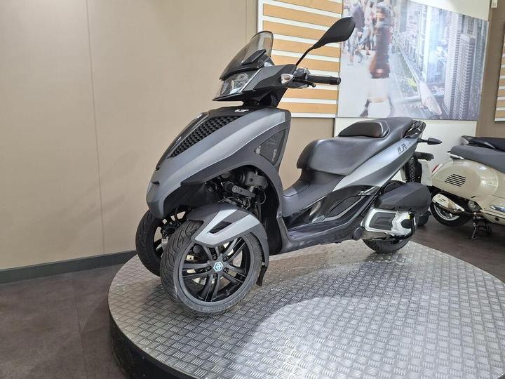 A sleek gray three-wheeled scooter is displayed on a circular platform, featuring a modern design with a front windshield, digital dashboard, and stylish alloy wheels. In the background, a blurred cityscape image is visible, along with another classic scooter.