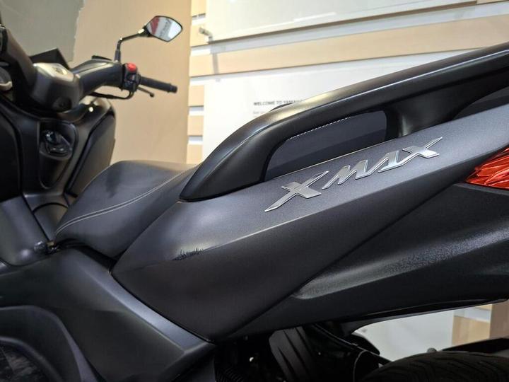 Close-up of the side panel of a black scooter with the model name "XMAX" prominently displayed, showcasing a sleek design and leather seat. In the background, a blurred interior with light colors is visible.