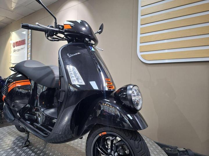 A black Vespa scooter with orange accents is parked in a showroom, featuring a sleek design and illuminated headlight, against a neutral background with wooden paneling.