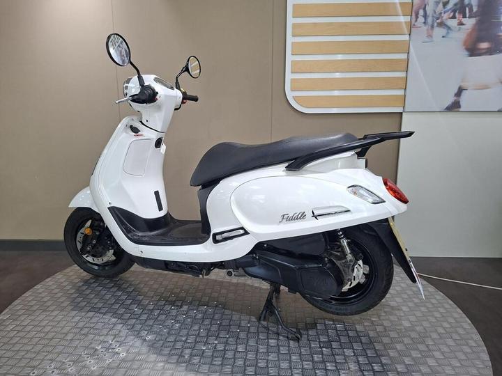 A side view of a white scooter parked on a textured surface, with a black seat and round mirrors. The scooter features a sleek design and the model name "Fiddle" visible on the rear. The background consists of a simple beige wall.