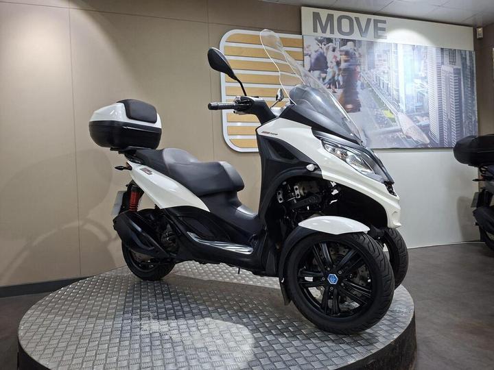 A stylish three-wheeled scooter in white with a black seat and back storage compartment, displayed on a circular metallic platform against a modern backdrop featuring a cityscape and the word "MOVE."