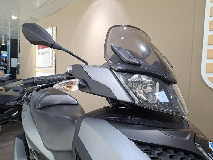 Close-up of a motorcycle's front, featuring the windshield, side mirror, and handlebar grips, with a blurred display of other motorcycles in the background.
