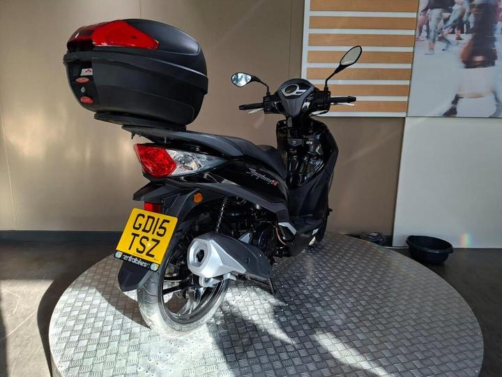 A black scooter with a top case is displayed on a circular textured platform, featuring a registration plate that reads "GD15 TSZ." The background has a neutral color and some blurred imagery.